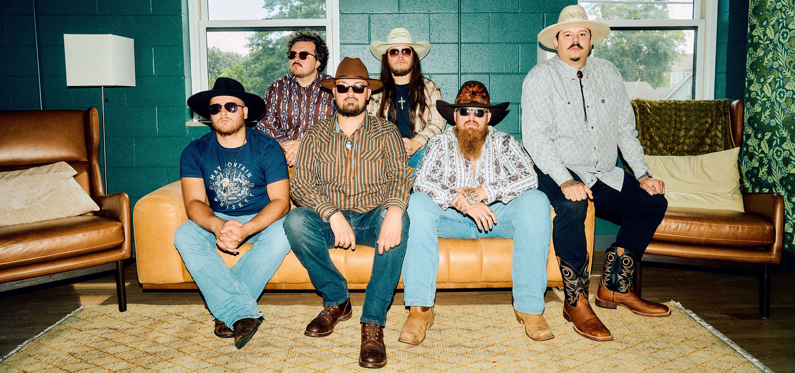 49 Winchester band members in hats and sunglasses seated on leather sofas in a retro lounge with teal walls and windows.