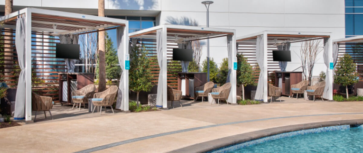 Row of modern poolside cabanas with TVs, lounge chairs, and slatted wood panels beside the pool.