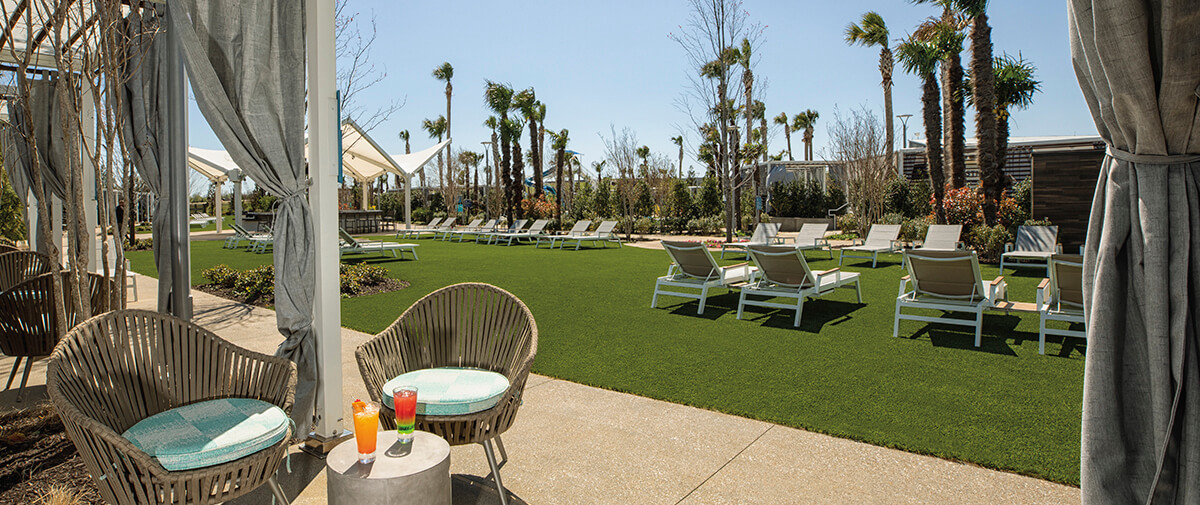 Grassy sun lawn lined with lounge chairs and palm trees, viewed from a shaded cabana seating area.