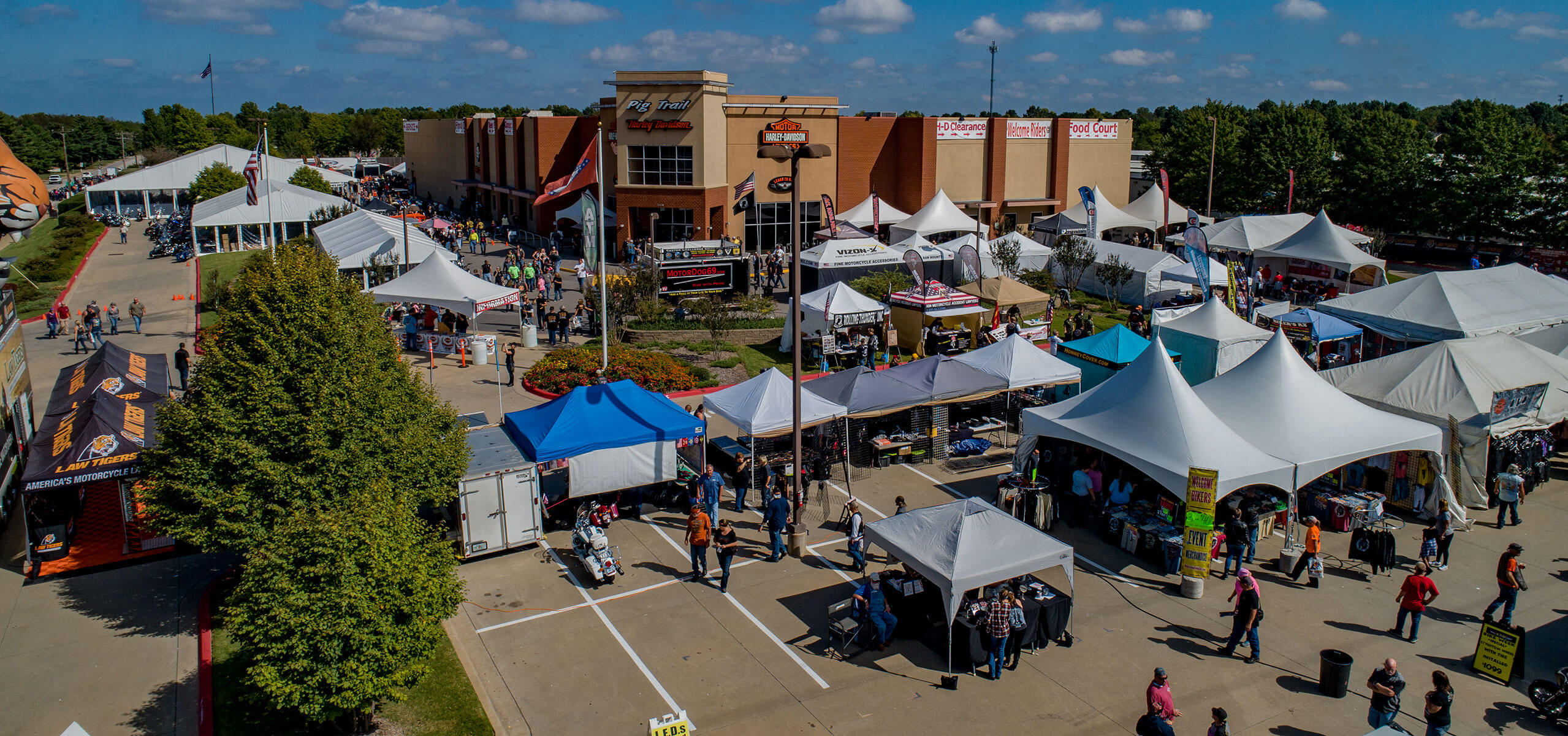 Drone view of an outdoor festival with rows of white vendor tents, crowds, and motorcycles outside a Harley-Davidson showroom on a sunny day