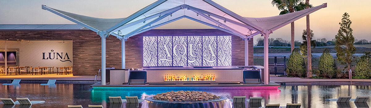 AQUA pool bar with sculptural canopy, glowing backlit ‘AQUA’ panel, in-pool loungers, and a fire feature at dusk; LUNA bar visible at left.