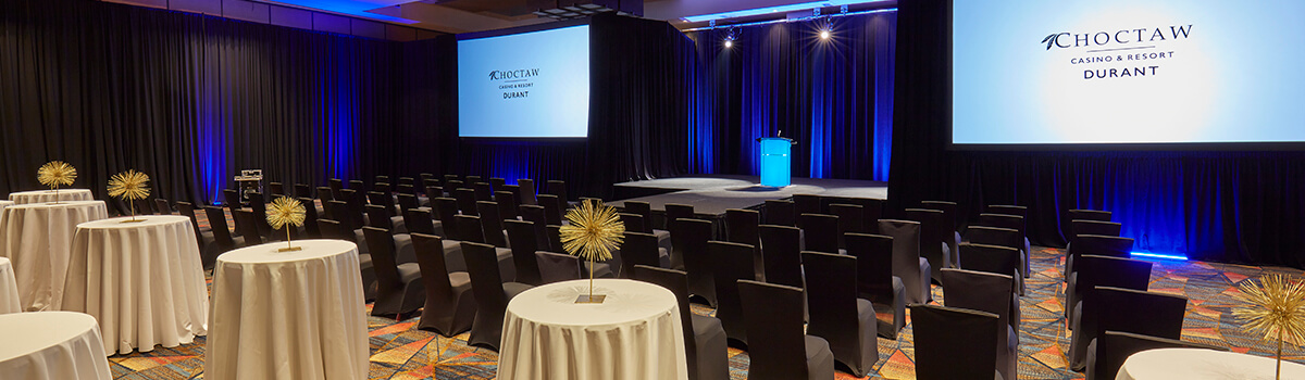 Conference room with rows of black chairs facing two ‘Choctaw Casino & Resort – Durant’ screens, blue uplighting, a podium, and round cocktail tables with gold starburst centerpieces
