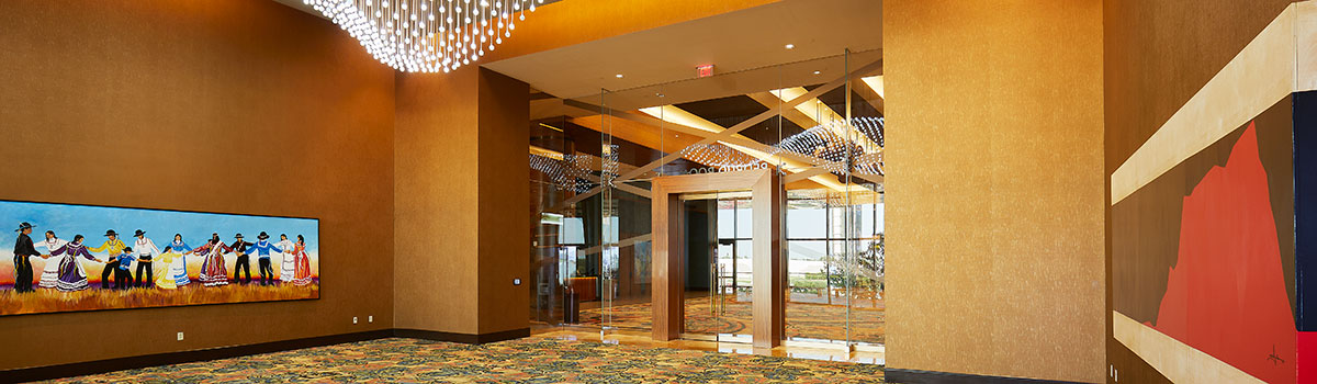 Warm lobby outside a meeting room with glass doors, modern chandeliers, patterned carpet, and Native art along the wall.