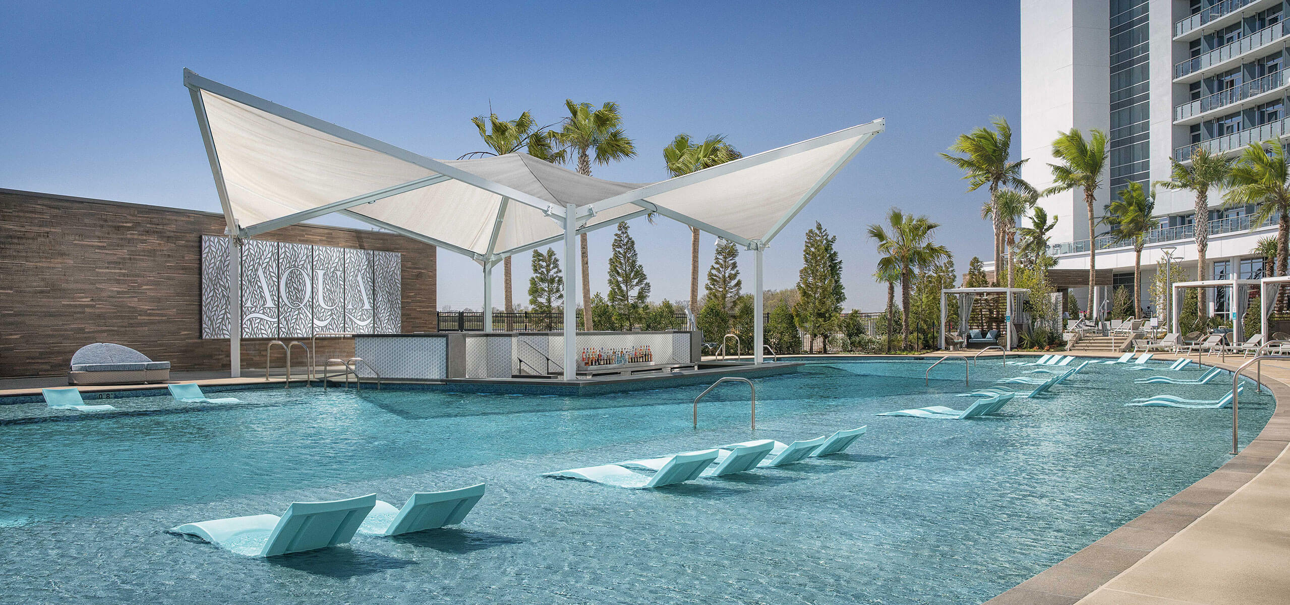 Aqua resort pool with white sail-like canopy, in-water loungers, palm trees, and the Choctaw Durant hotel tower in the background