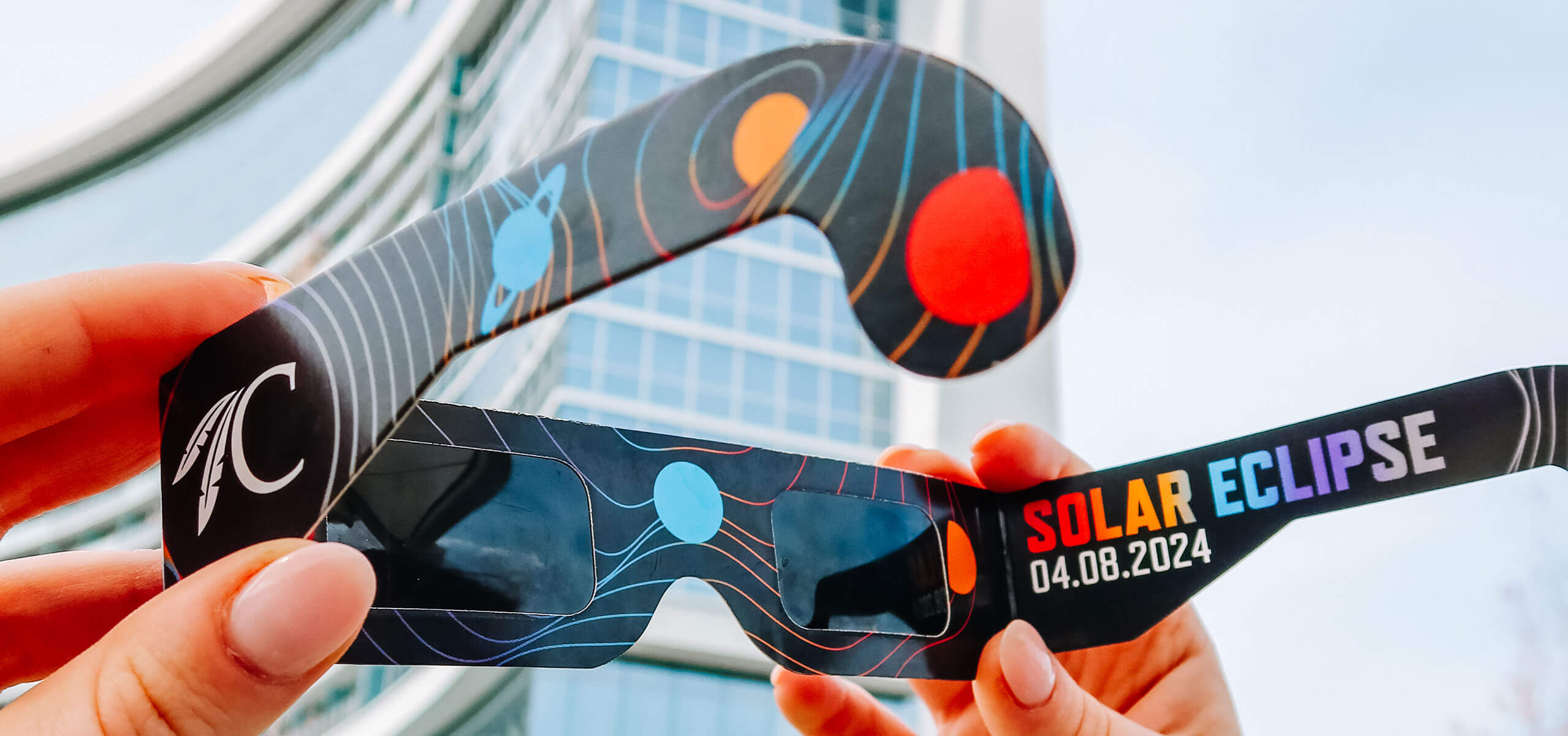 Hands holding branded eclipse glasses that read ‘SOLAR ECLIPSE 04.08.2024’ in front of a Choctaw hotel building