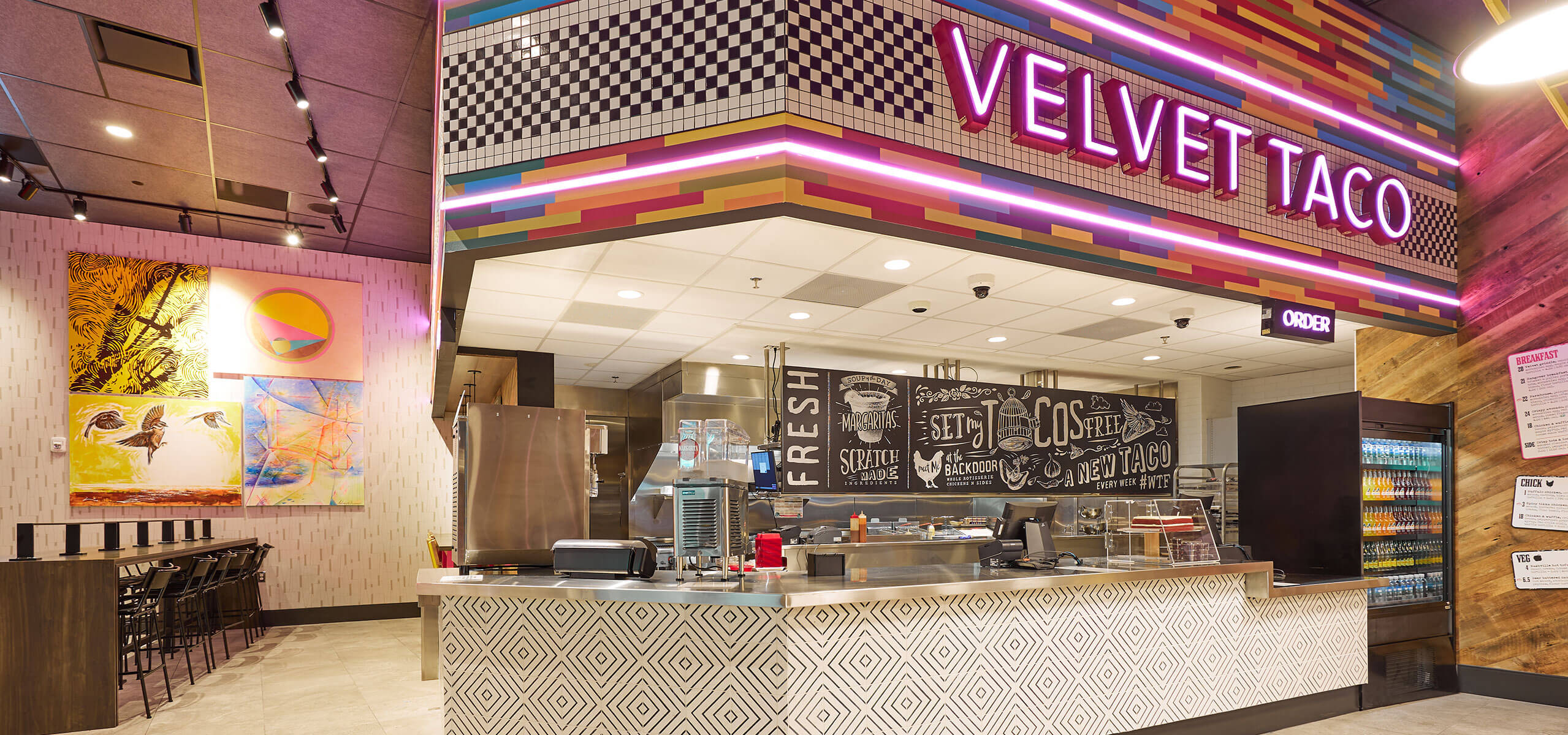 Velvet Taco counter at the District Food Hall with pink neon sign, colorful tile soffit, open kitchen, and order counter.