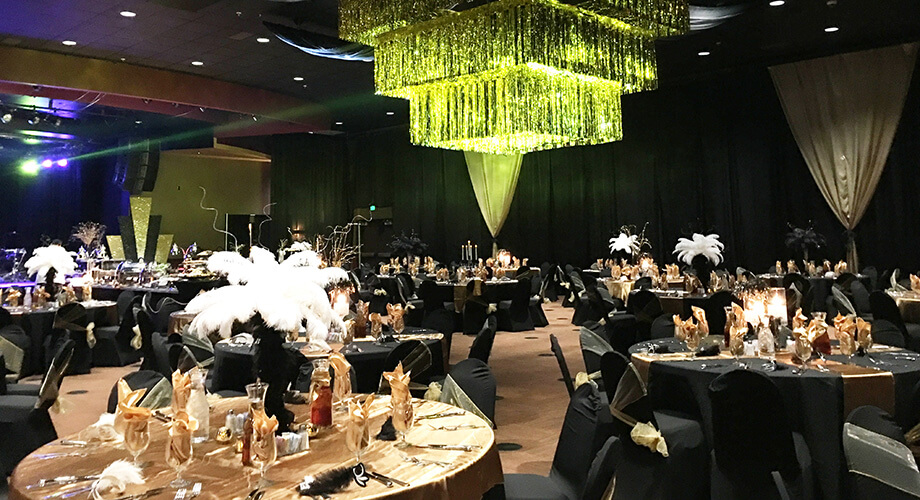 Center Stage event space set for a gala with black tablecloths, gold accents, feather centerpieces, and a hanging gold-tinsel chandelier.