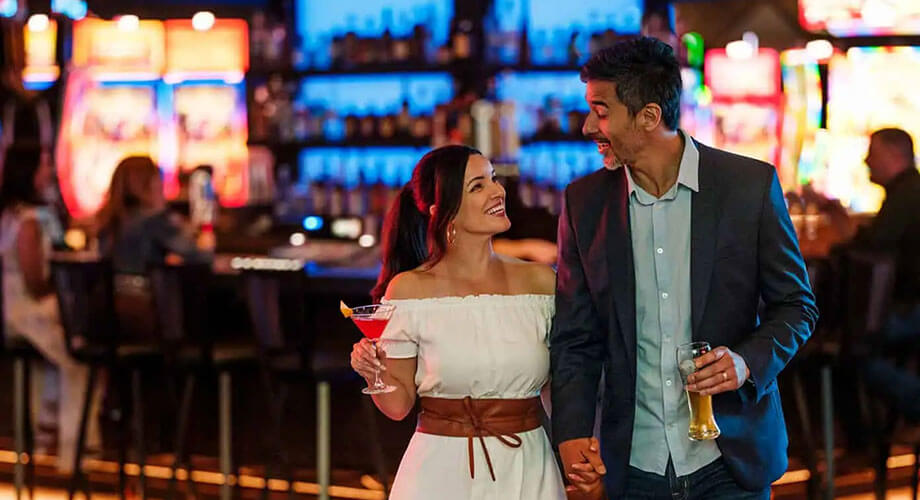 Two guests holding drinks and smiling as they walk past a glowing center bar with slot machines in the background