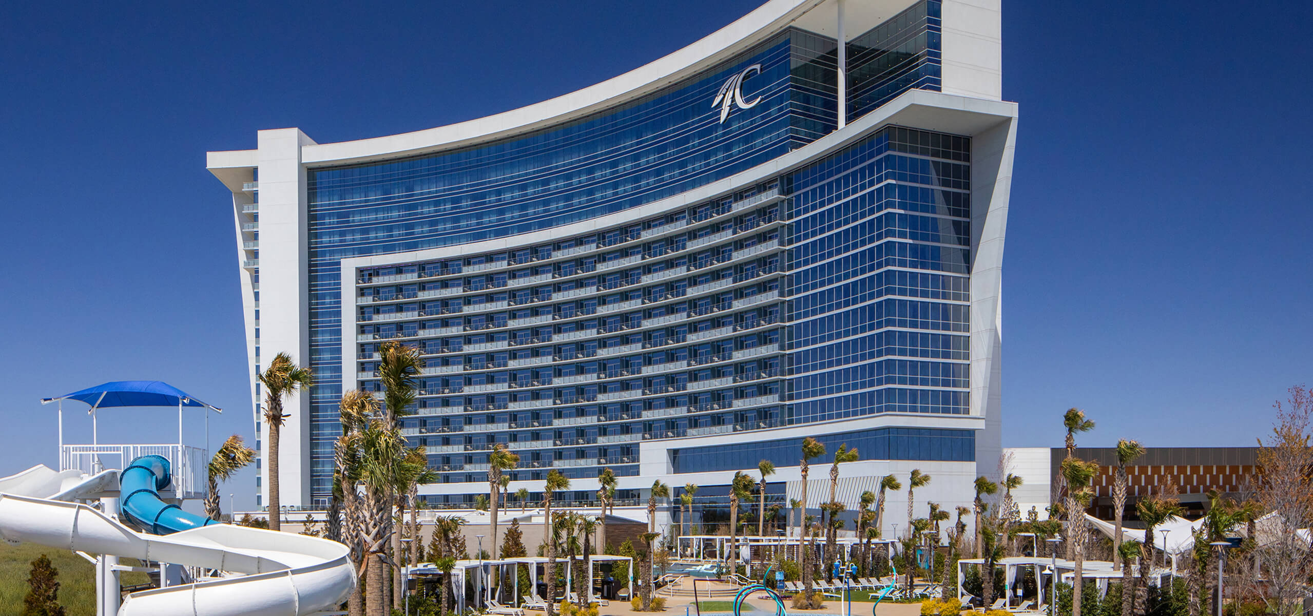 Curved glass Durant hotel tower overlooking the AQUA pool area with waterslide, palms, cabanas, and a clear blue sky.